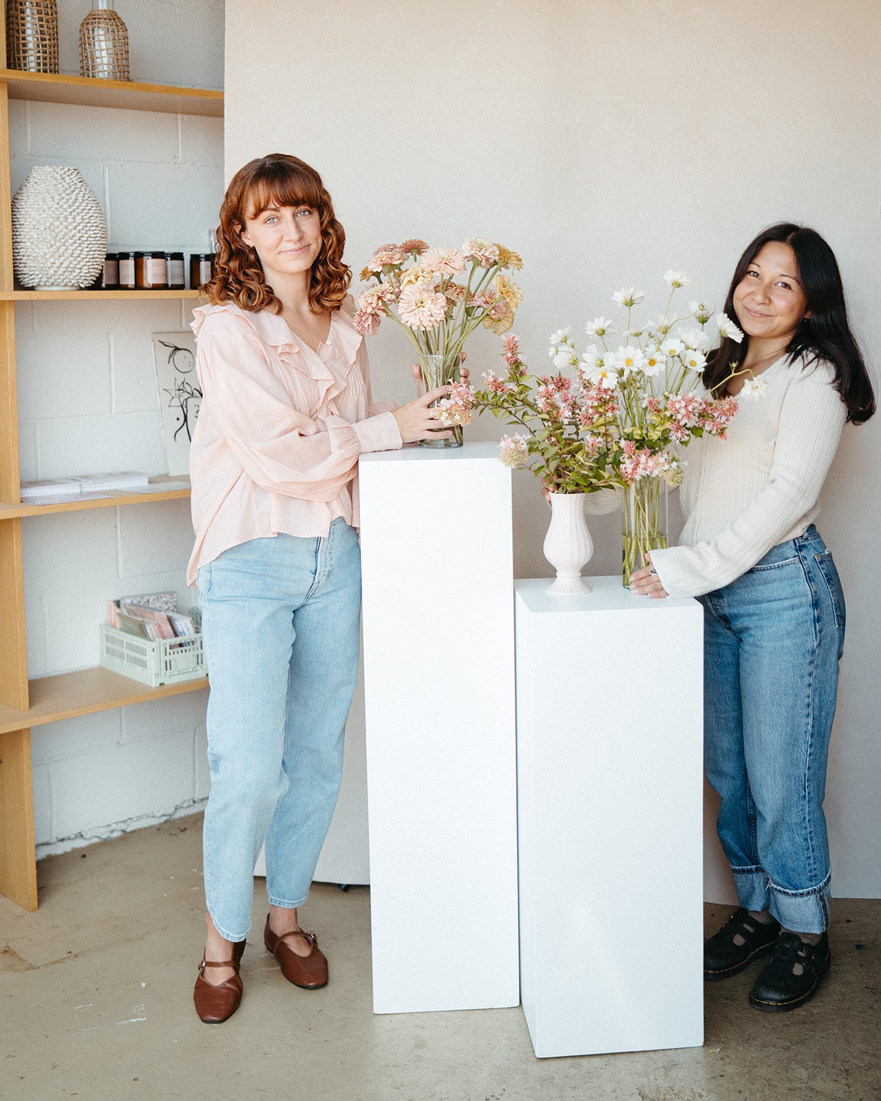 Two women standing in a room with shelves and flowers, one holding a bouquet.